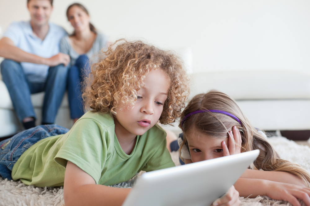 Young children using a tablet computer while their happy parents are watching in their living room Young children using a tablet computer while their happy parents are watching in their living room